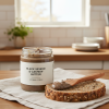 A jar labeled “Black Sesame & Cashew” rests on a cloth as a wooden knife spreads the butter over multigrain bread in a kitchen setting.