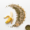 A peeled banana beside scattered pumpkin seeds, sunflower seeds, and flaxseeds on a white surface, with two small wooden bowls holding seeds.