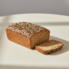 A loaf of seeded bread with one slice cut off sits on a plain surface, topped with a variety of seeds.